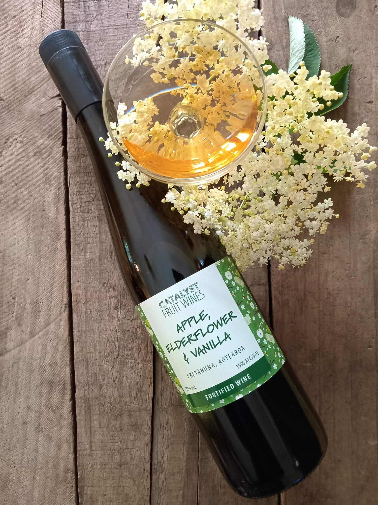 Bottle of Catalyst Fruit Wines with a glass of wine and elderflower on a wooden surface